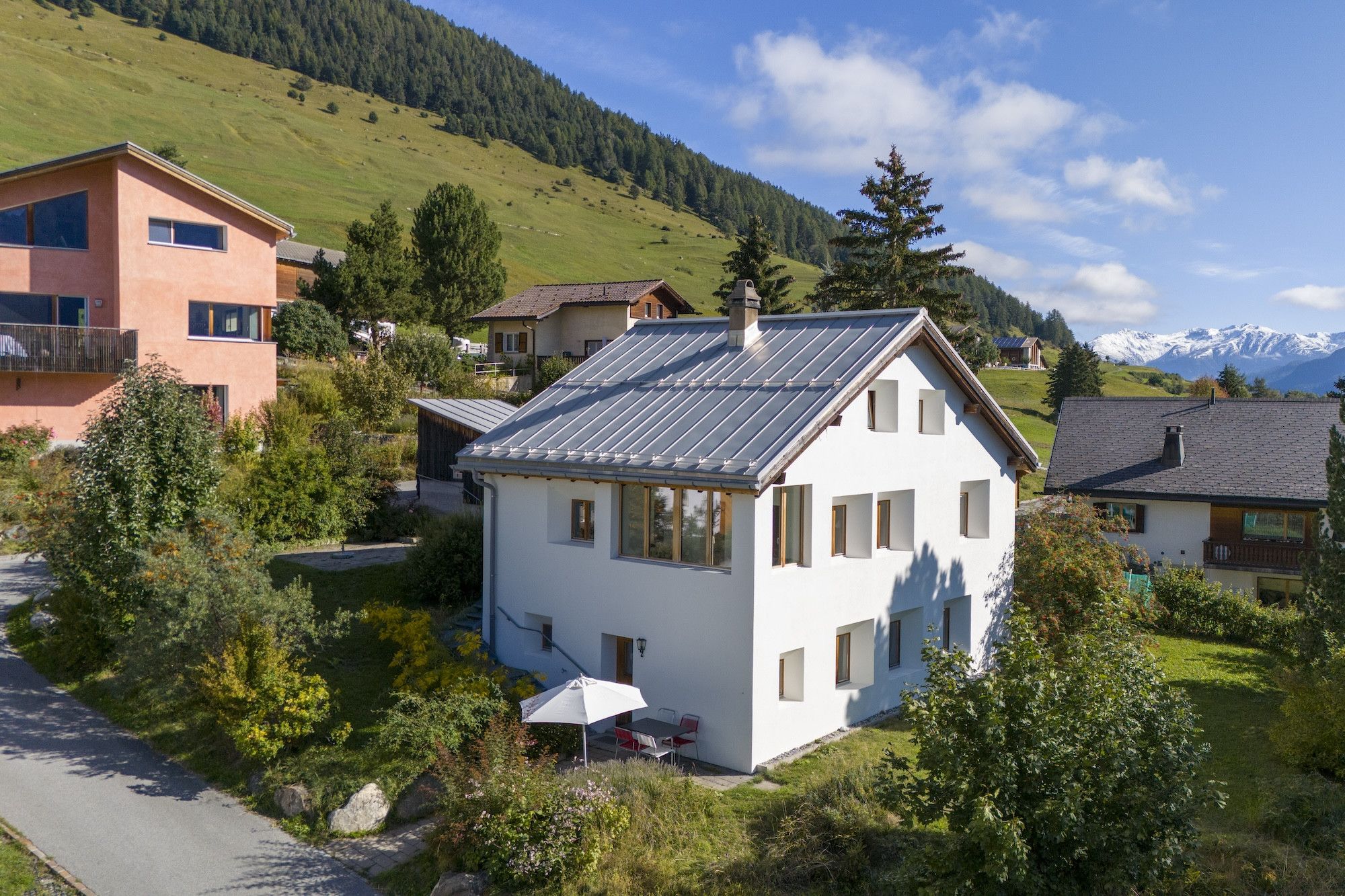 A white house with a sloped roof, surrounded by lush greenery and a mountainous backdrop.