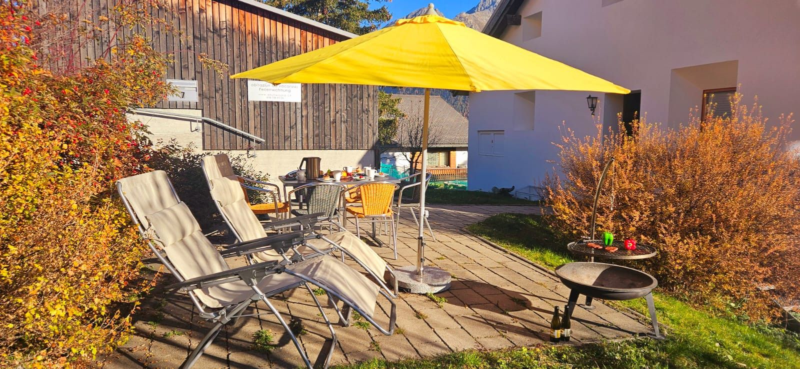 Un patio al aire libre con sillones de descanso, un paraguas amarillo, una mesa con bebidas y una parrilla.
