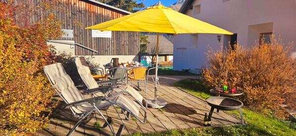 Un patio al aire libre con sillones de descanso, un paraguas amarillo, una mesa con bebidas y una parrilla.