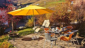 Une terrasse extérieure avec une table et des chaises, un parasol jaune et une vue panoramique sur des collines et des maisons.