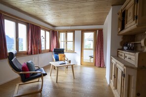 A room with a wooden ceiling, a desk, a chair with cushions, a window with curtains, and a wooden cabinet.