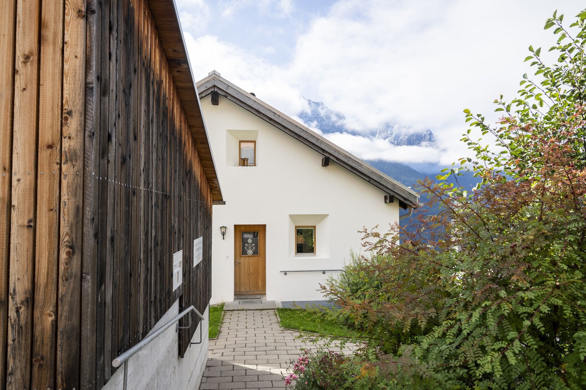 Une maison à deux étages, avec une façade blanche et un revêtement extérieur en bois, dotée d’un petit porche à l’avant et surplombant un paysage montagneux.