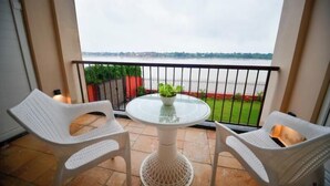 En balkon med to hvide fletstole, et glasbord med en grøn plante og udsigt til en flod og grøntområde.