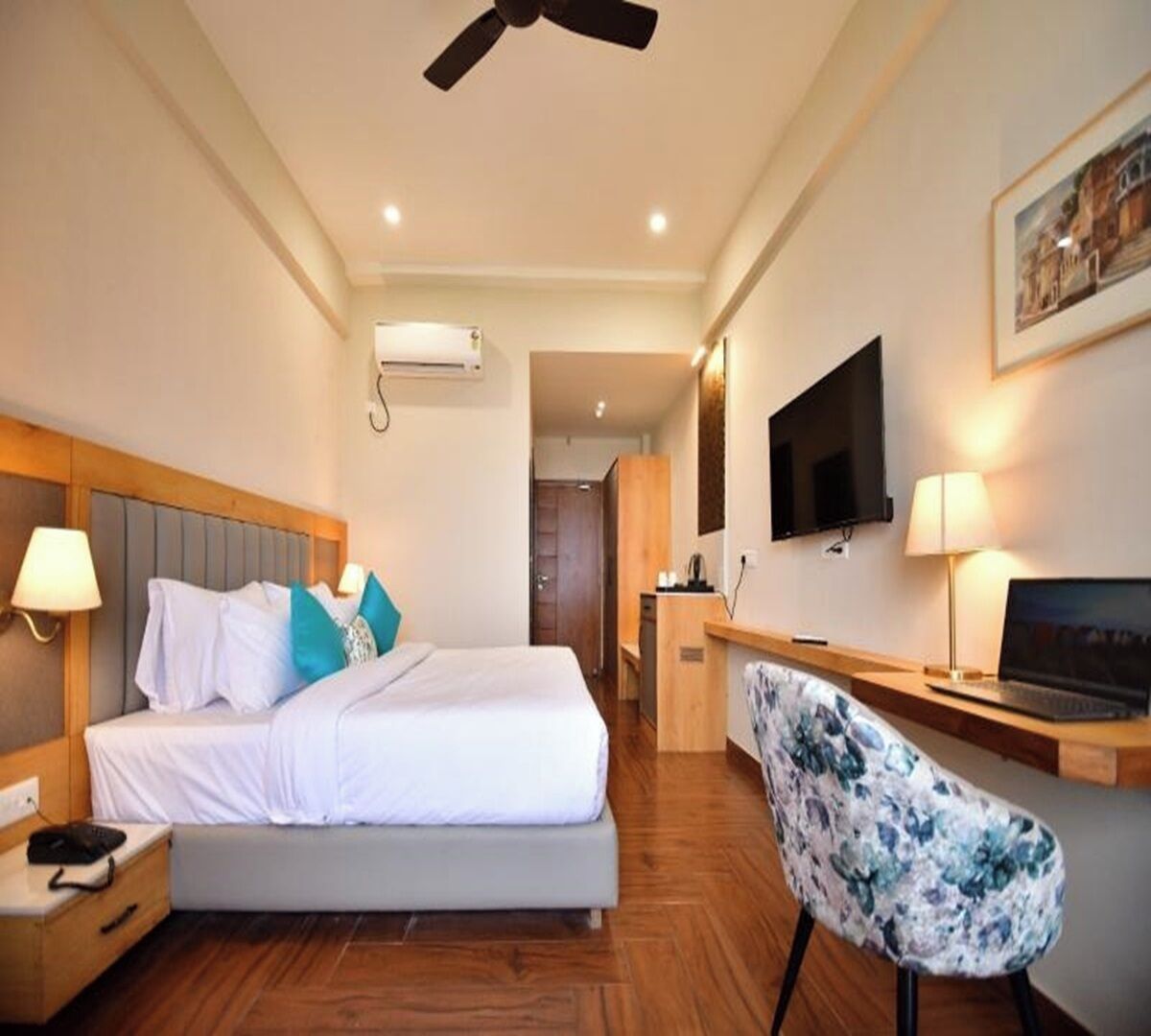 A hotel room with a bed, bedside lamps, a desk with a laptop, a chair, a TV, and an air conditioning unit.
