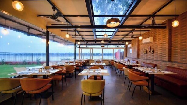 En restaurant med højt til loftet, synlige træbjælker og udsigt til det fri. Borde og stole er sat frem til spisning.