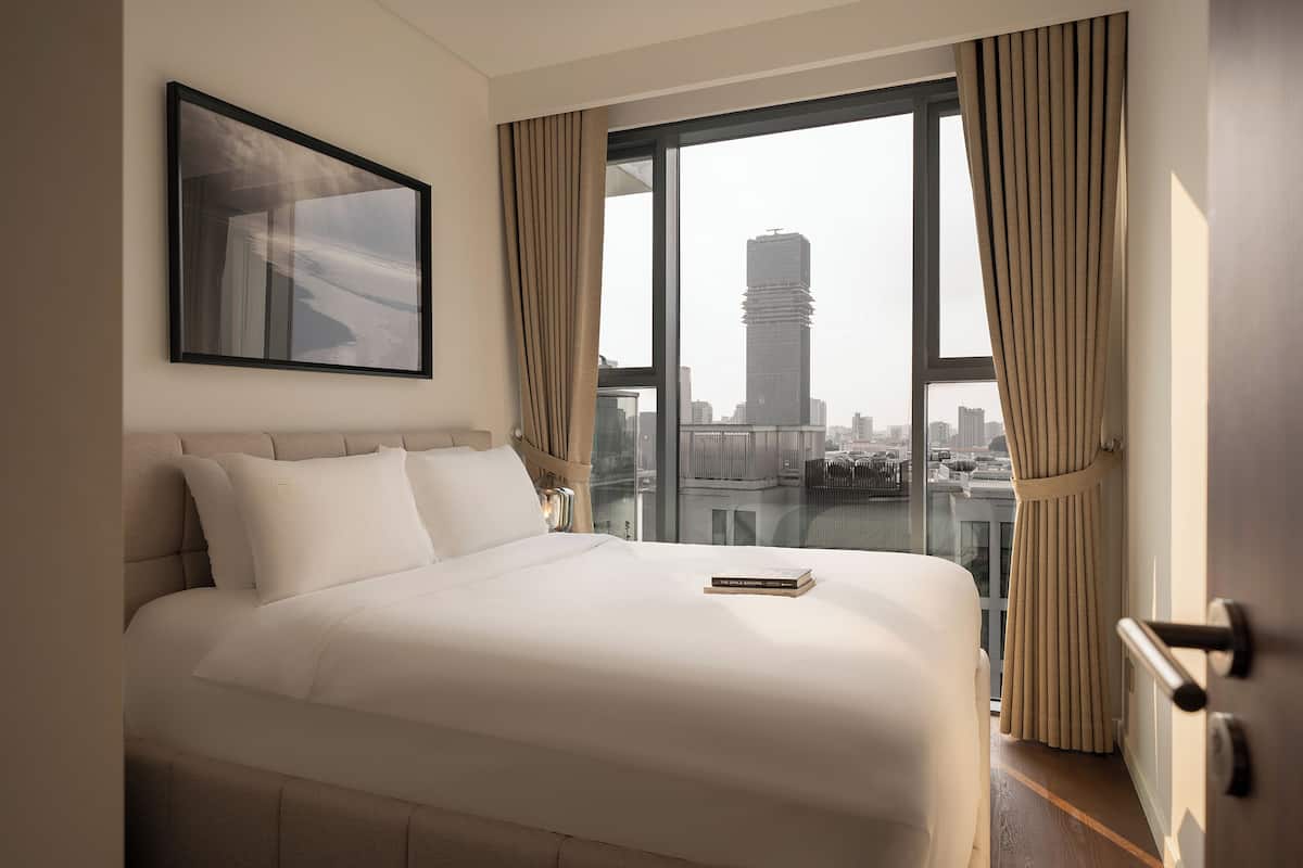 Ein Hotelzimmer mit einem großen Bett, einem gerahmten Bild, Blick auf eine Stadtlandschaft durch ein Fenster und einer Tür mit Griff.