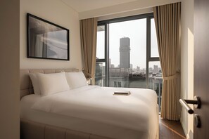 A hotel room with a large bed, a framed picture, a view of a cityscape through a window, and a door with a handle. - The Metropole Signature Residence by CR Concept (Ho Chi Minh City)
