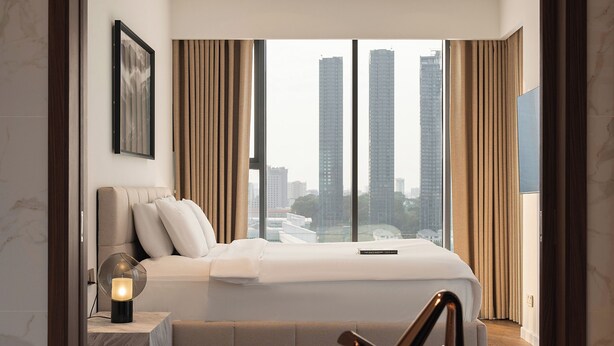 Een moderne slaapkamer met een groot bed, een nachtkastje, een bijzettafeltje met een lamp, uitzicht op hoge gebouwen en een badkuip.