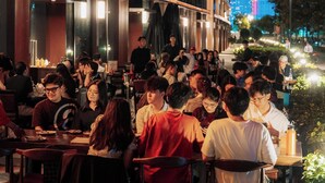 Een terras met tafels en stoelen, omgeven door een gebouw met grote ramen en uitzicht op een verlicht stadsbeeld.