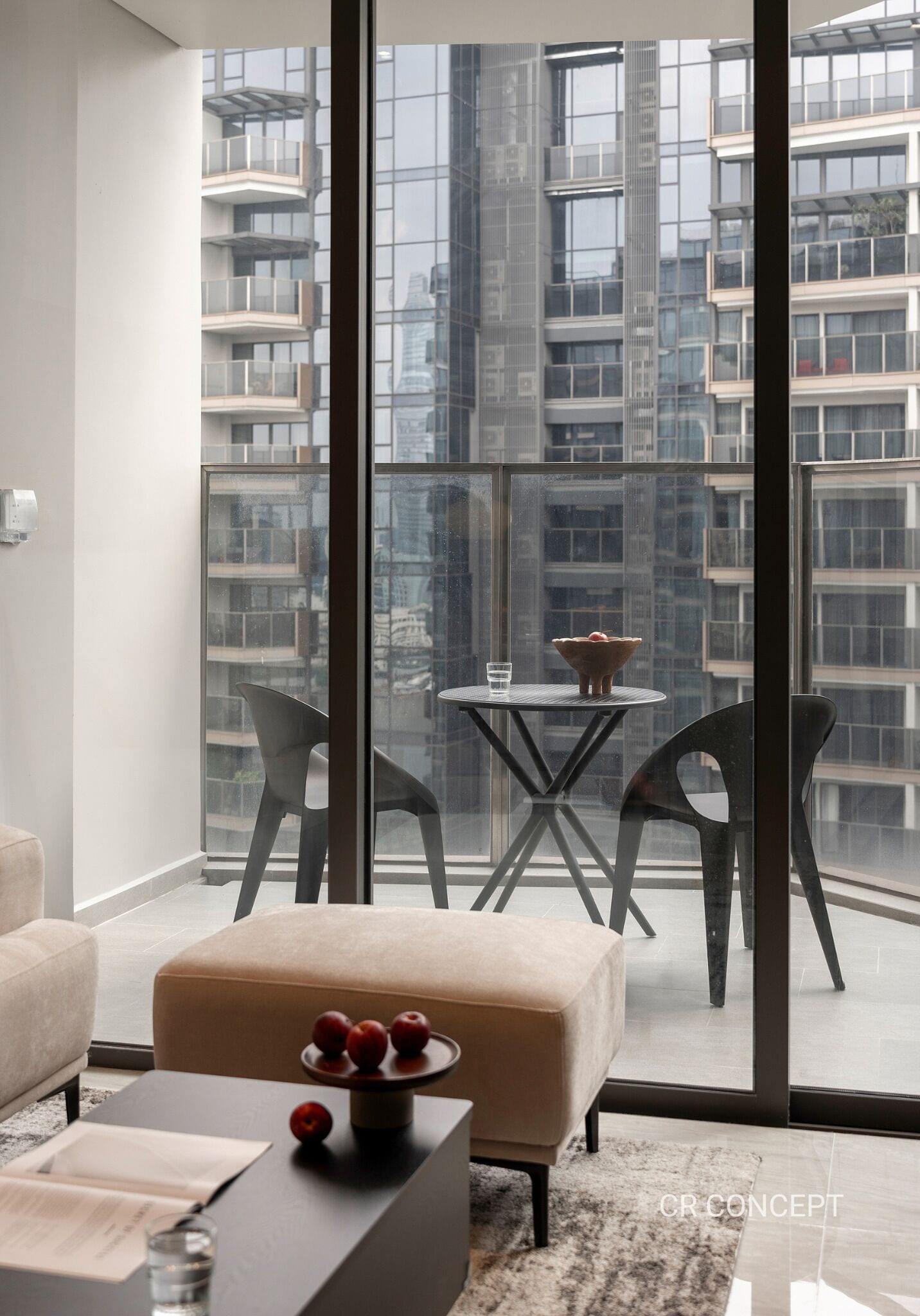 A modern living room with a large glass door leading to a balcony, a round table with two chairs, and a sofa with a small table and a bowl of fruit.