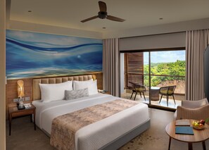 A bedroom with a large bed, a ceiling fan, a nightstand, a window with a view of greenery, and a seating area with a table and chairs. - Gateway Goa, Palolem (Canacona)