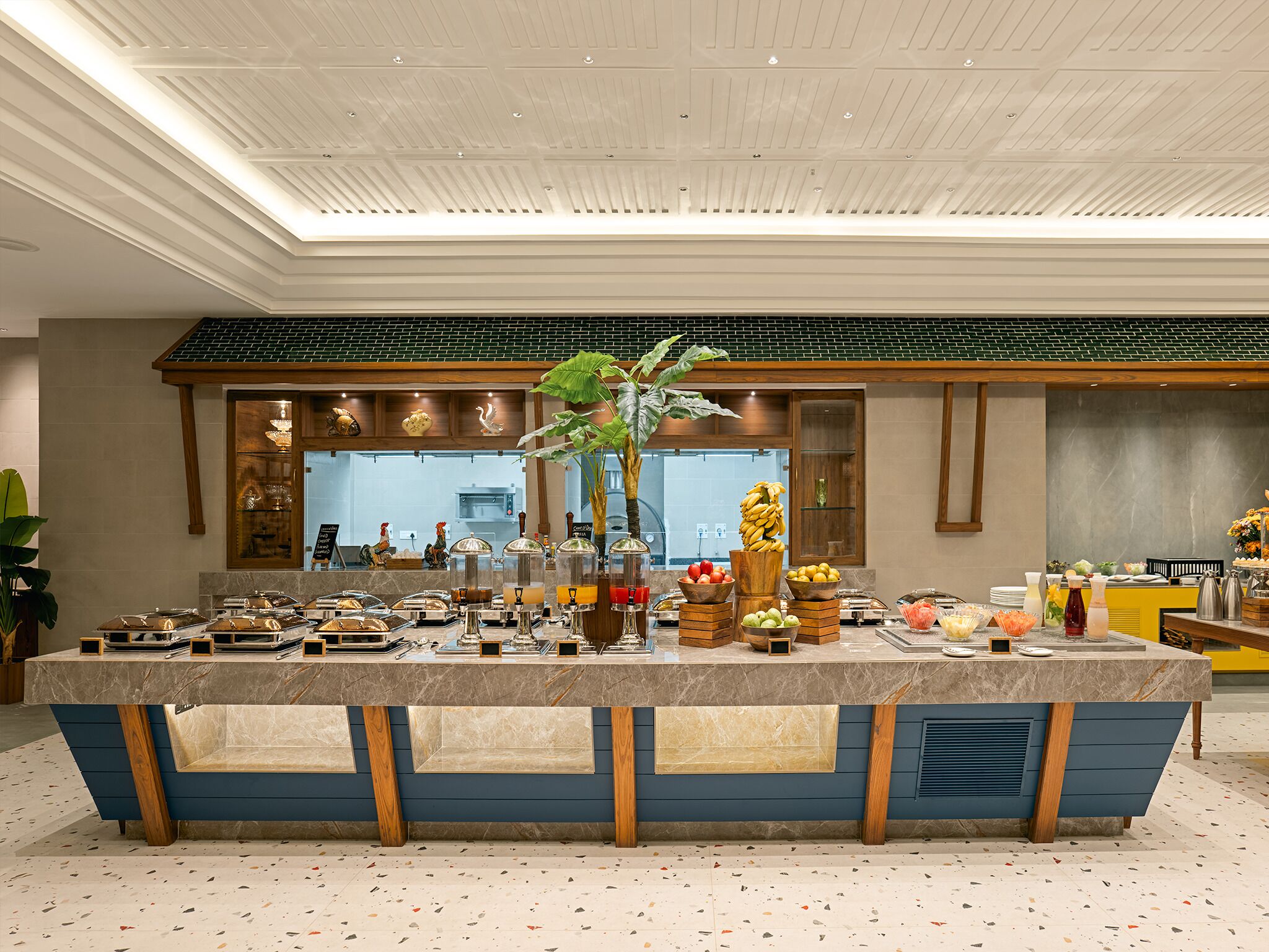 A breakfast buffet area with a central island, various food items, and a kitchen in the background.