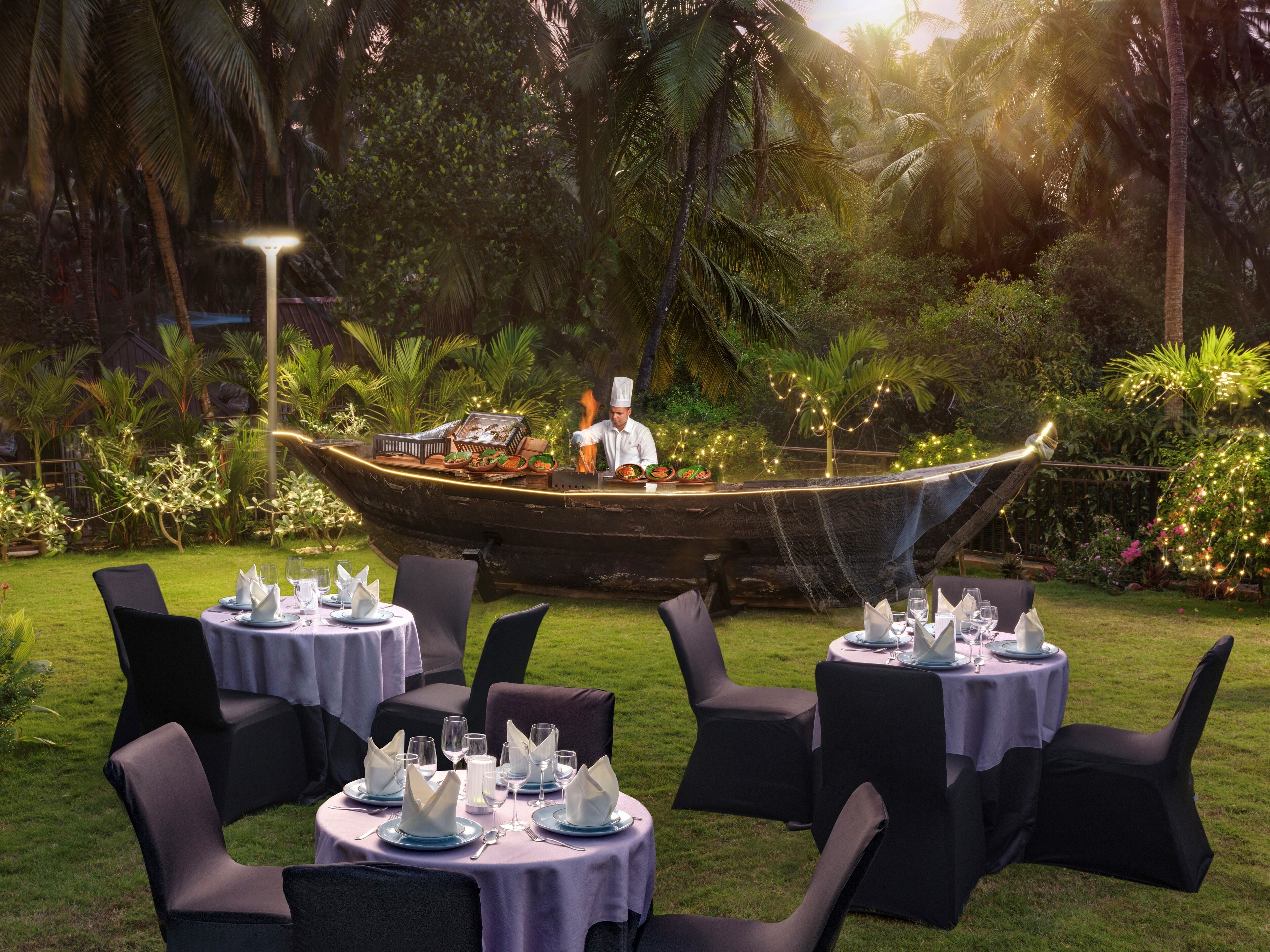 An outdoor dining area with tables set for a meal, a chef preparing food on a boat, and lush greenery in the background.