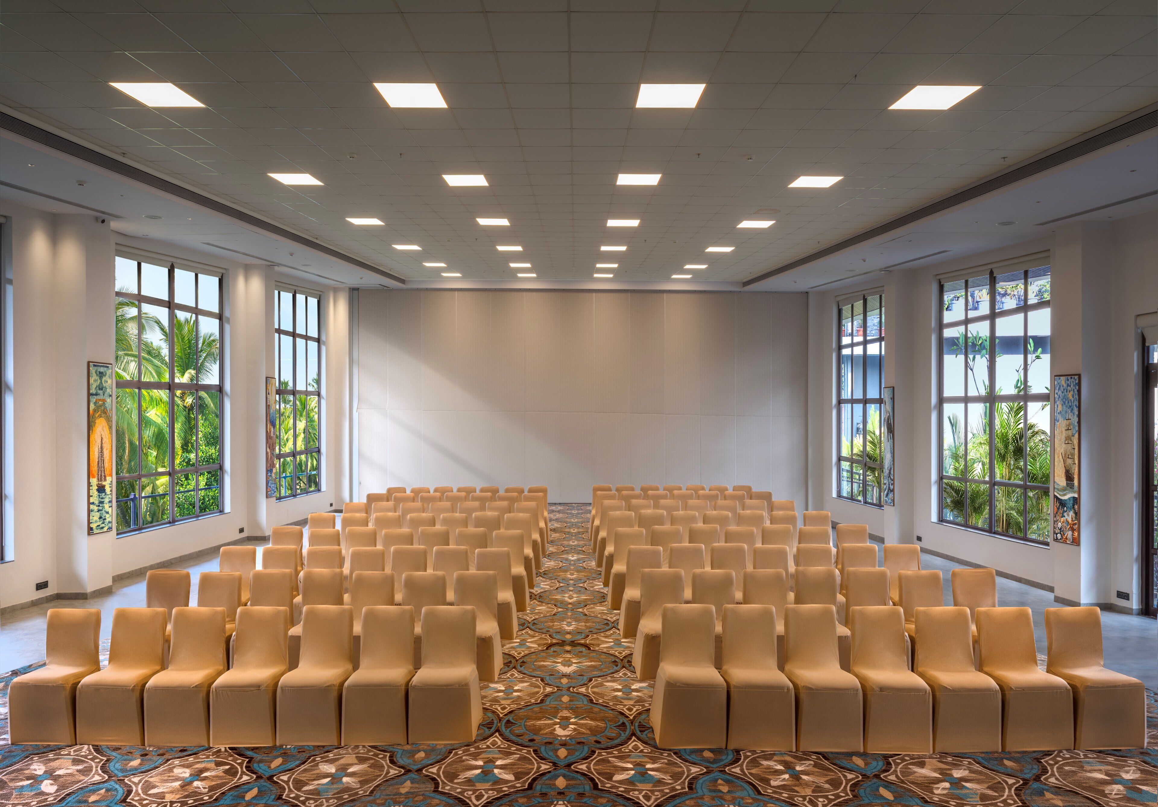 A conference room with rows of chairs, large windows with views of greenery, and a patterned carpet.