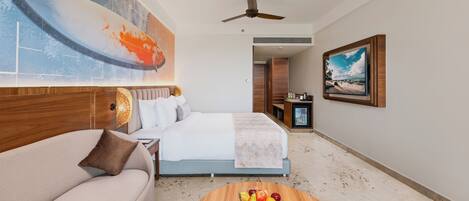 A hotel room with a bed, a sofa, a coffee table with fruit, a painting, and a ceiling fan.