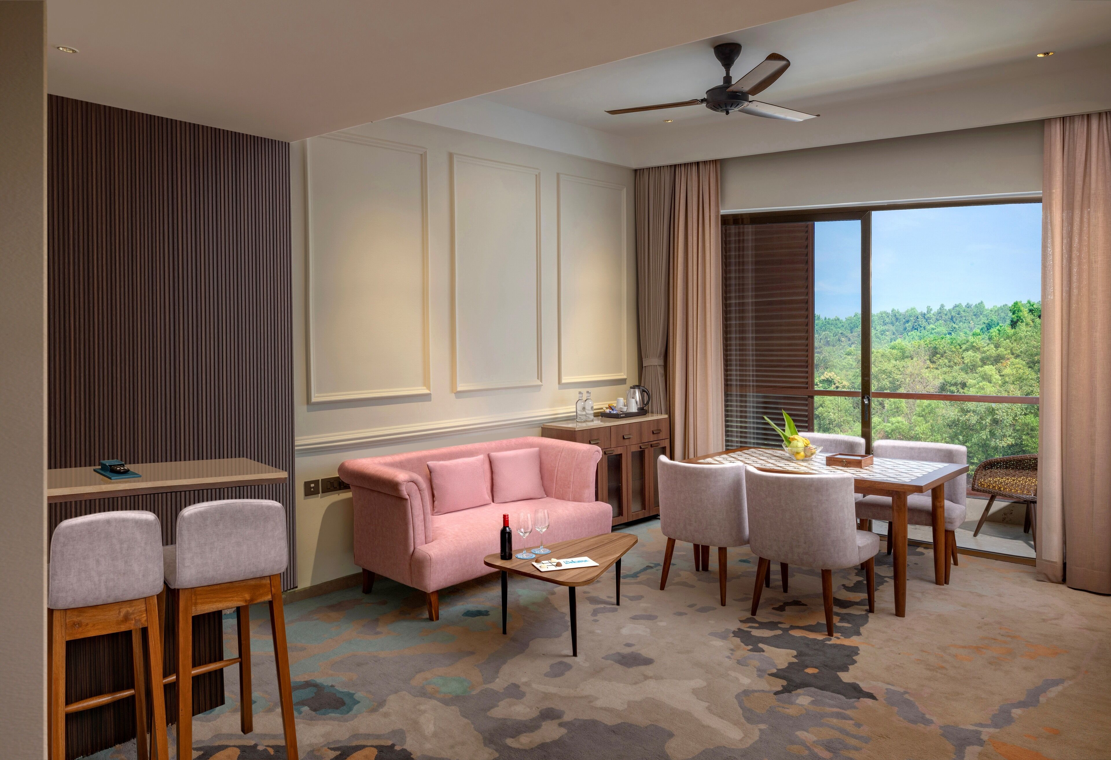 Habitación de hotel con sofá rosa, mesa de comedor con sillas, ventilador de techo y ventana grande con vistas a árboles.