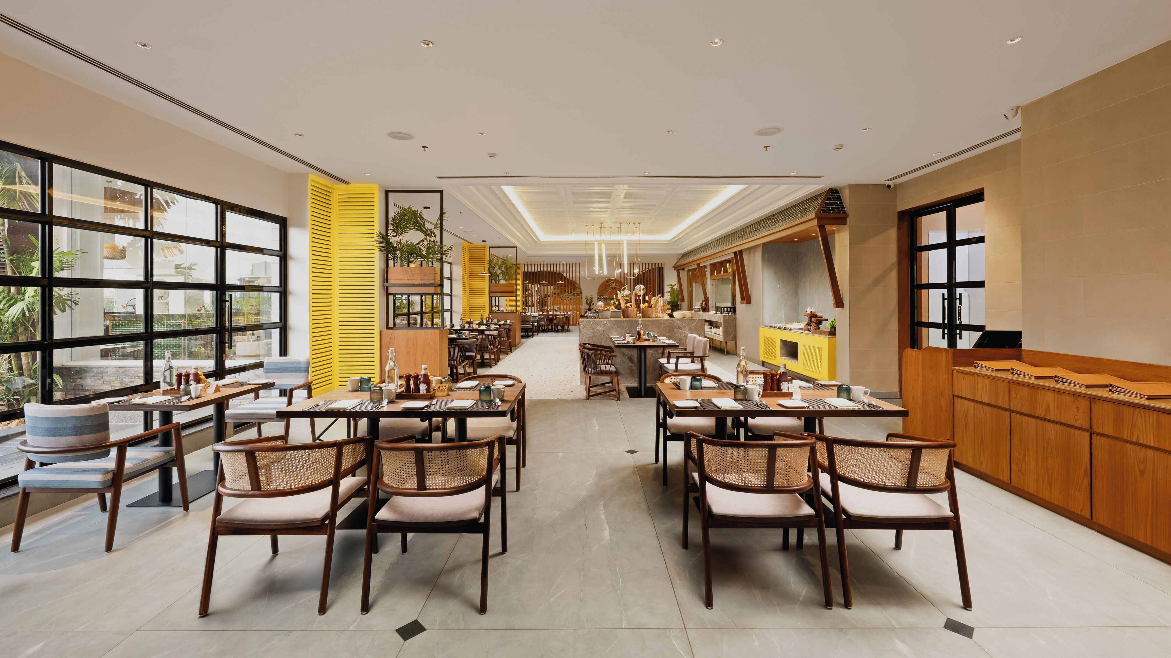 A spacious restaurant with high ceilings, large windows, and a central walkway. Tables are arranged on both sides, with chairs and a variety of table settings.