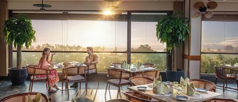 A restaurant with tables set for dining, large windows with a view, and hanging ceiling fans.