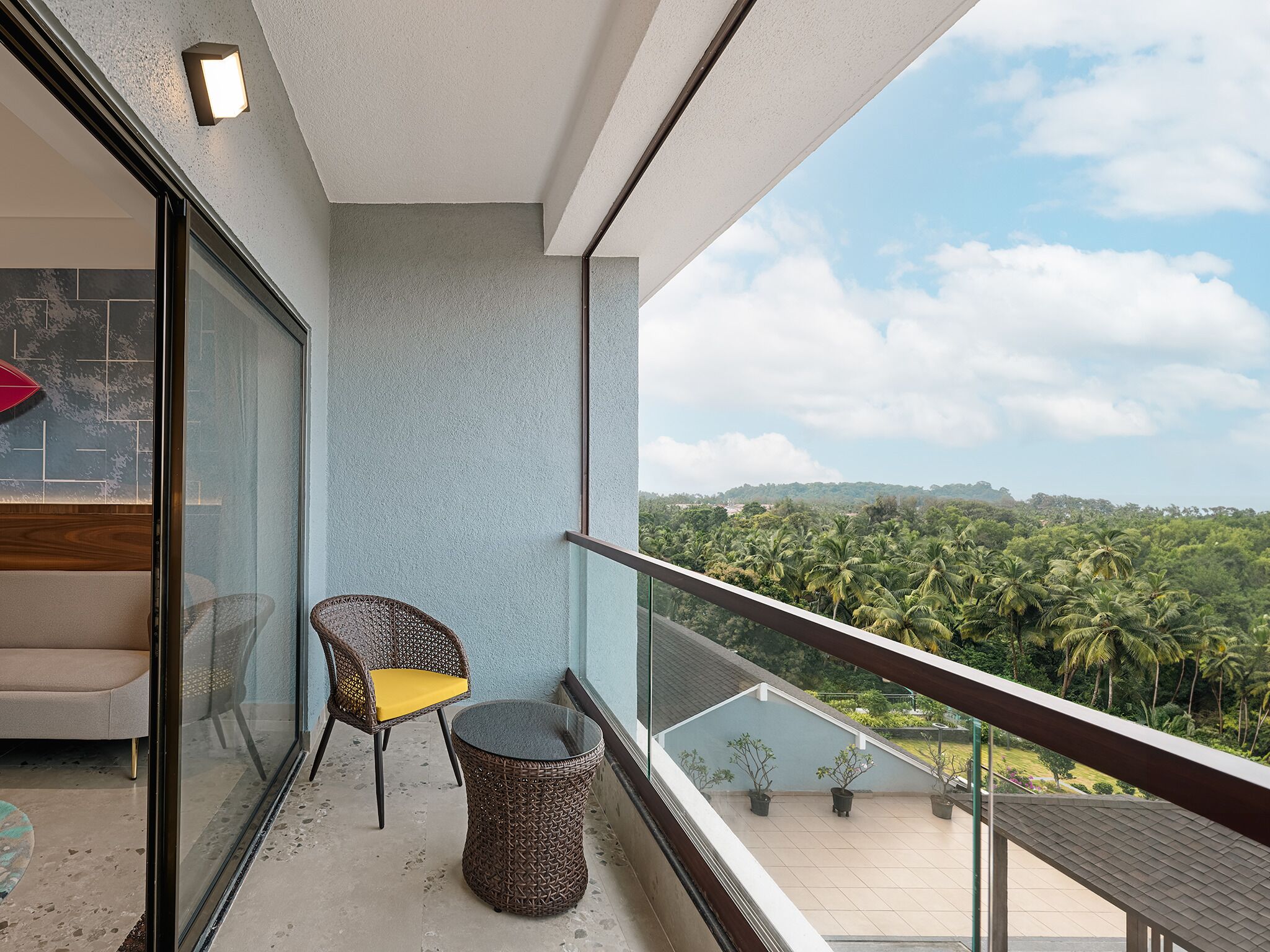 A balcony with a glass partition, a wicker chair with a yellow cushion, and a small round table.