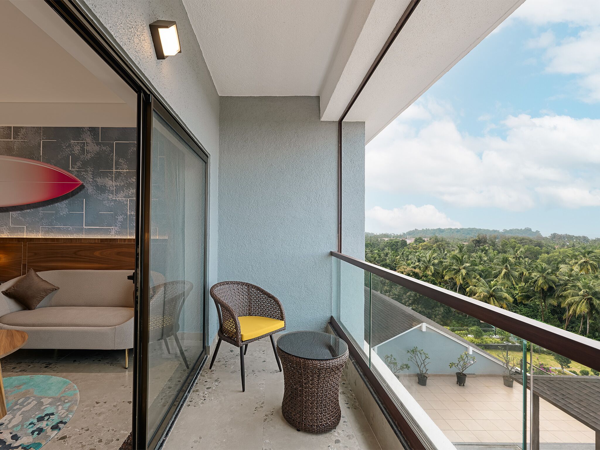 Ein Balkon mit Blick auf eine grüne Landschaft, ein Korbstuhl, ein kleiner Tisch und eine Couch mit Kissen.