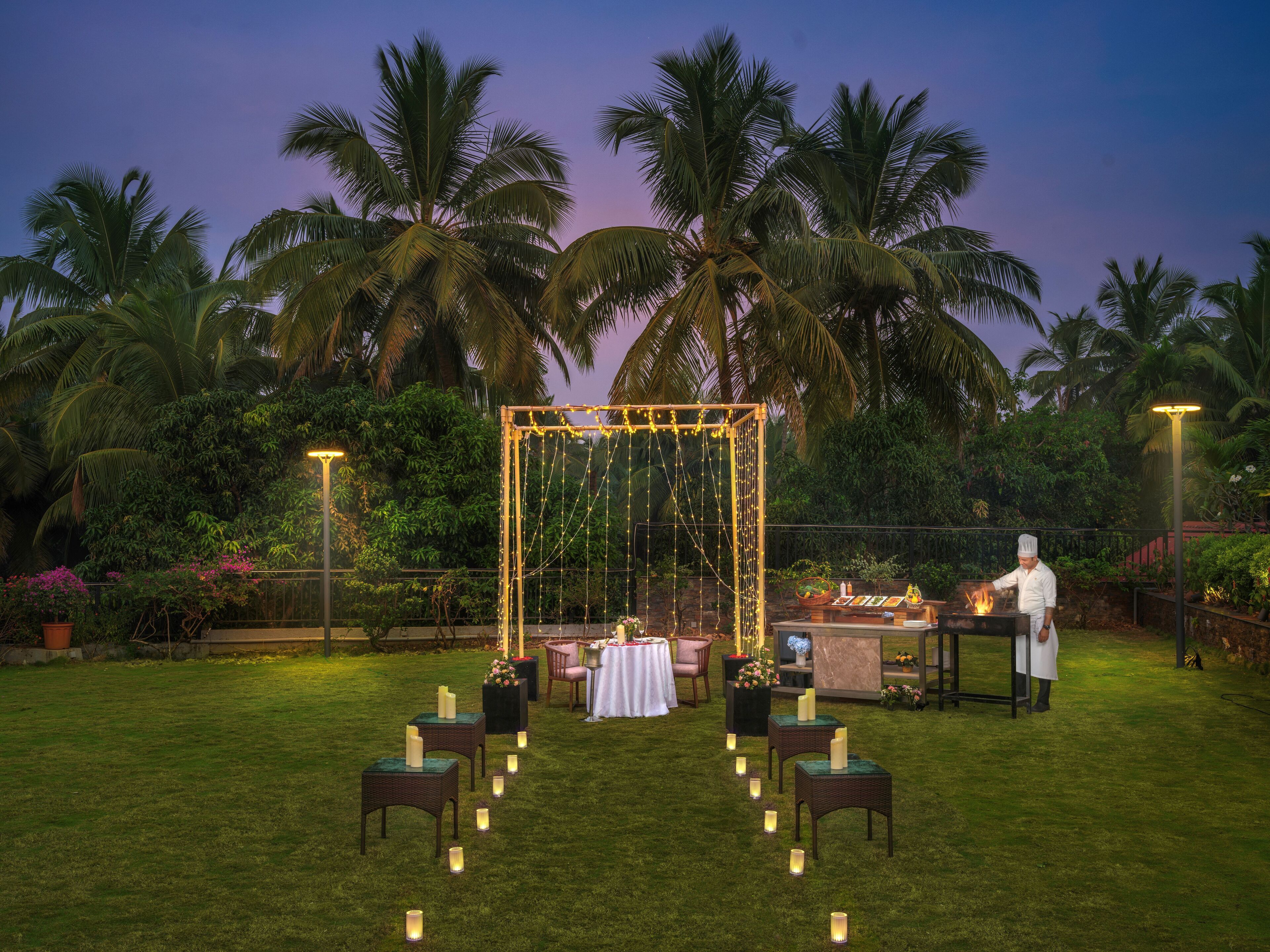 An outdoor wedding setup with a decorated arch, tables, chairs, and a chef preparing food.
