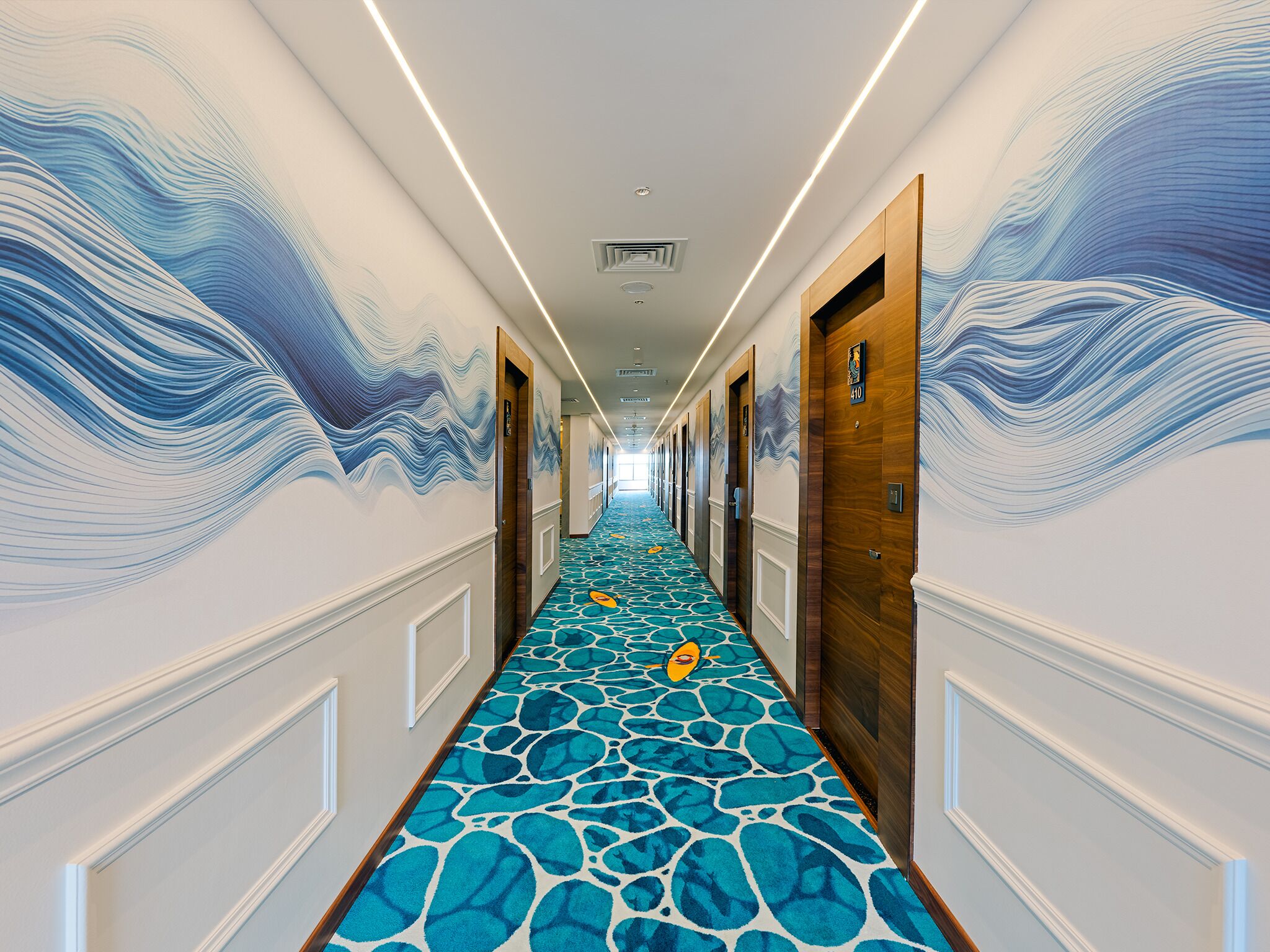 Un pasillo de hotel con alfombra estampada, puertas de madera y decoración de pared con motivos de olas.