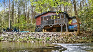 A rustic cabin with a deck and a staircase leading to an upper level, situated by a flowing river in a forested area. - Creekside Adventure! 10 steps from the cascading and gentle Fightingtown Creek (Blue Ridge)