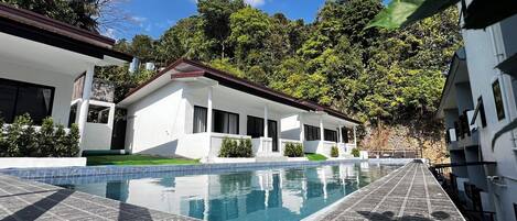 A modern building with a swimming pool, surrounded by lush greenery and a clear sky.