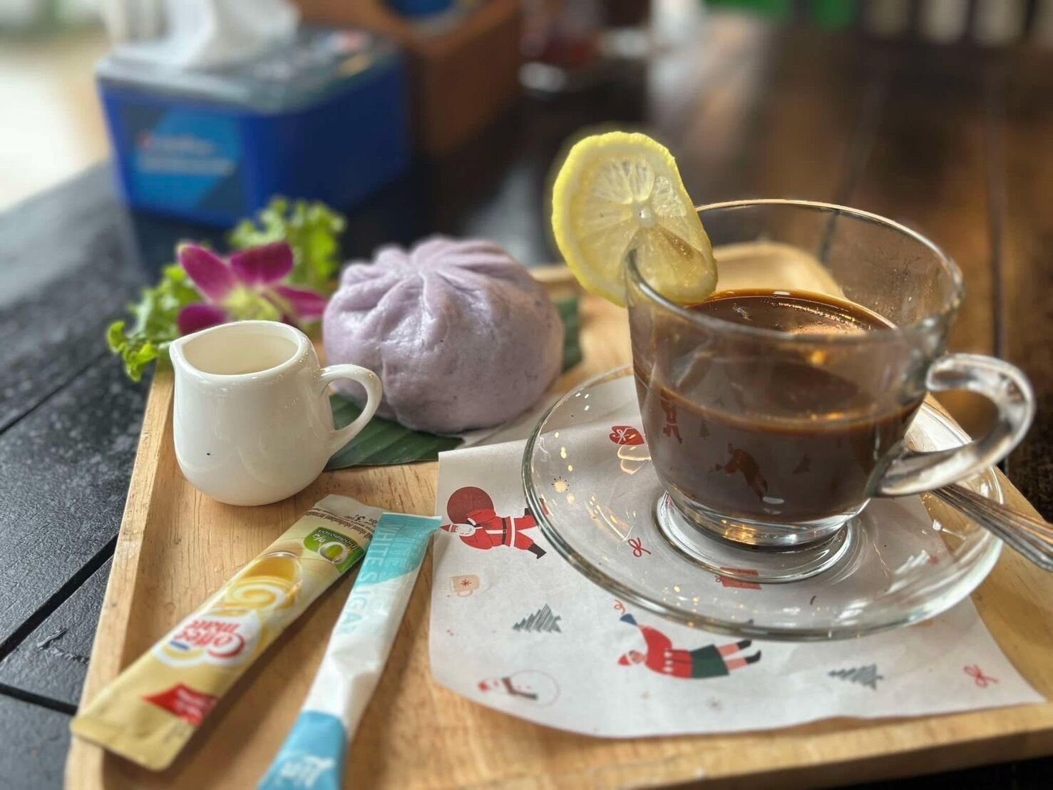 Una mesa con una taza de café, una taza blanca, un sobre de azúcar, un alimento morado y un paquete de té.