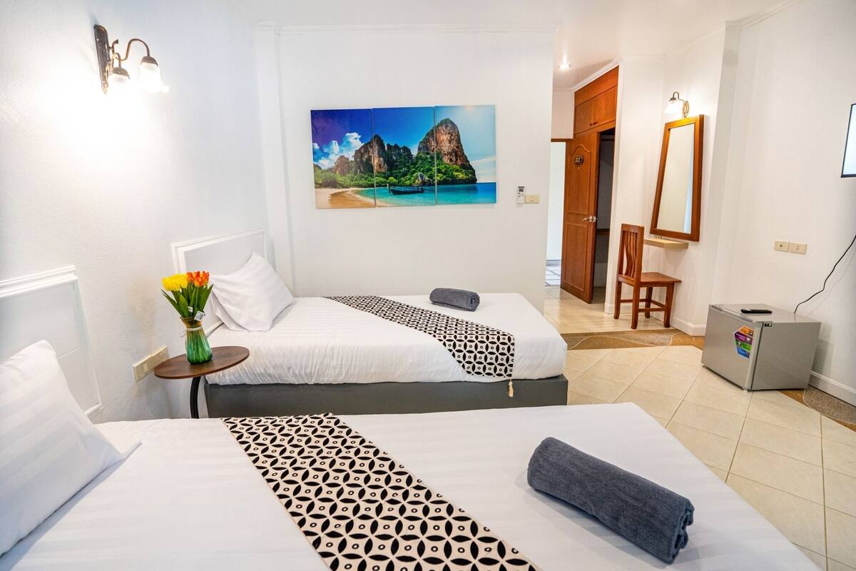 Ein Hotelzimmer mit zwei Betten, einem Gemälde von einem tropischen Strand, einem kleinen Tisch mit Blumenvase und einem Schreibtisch mit Computer.