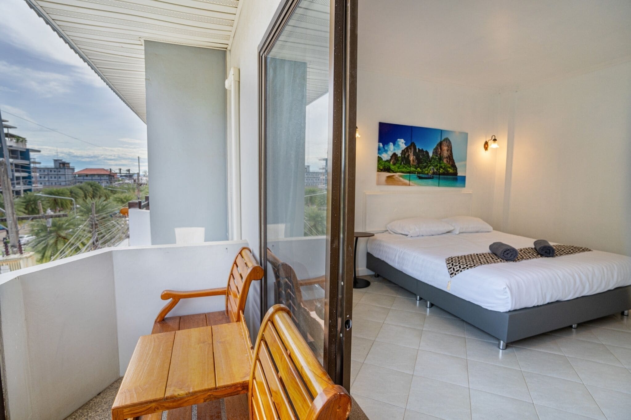A balcony with a wooden table and chairs, a bed with white bedding, and a painting of a coastal scene.