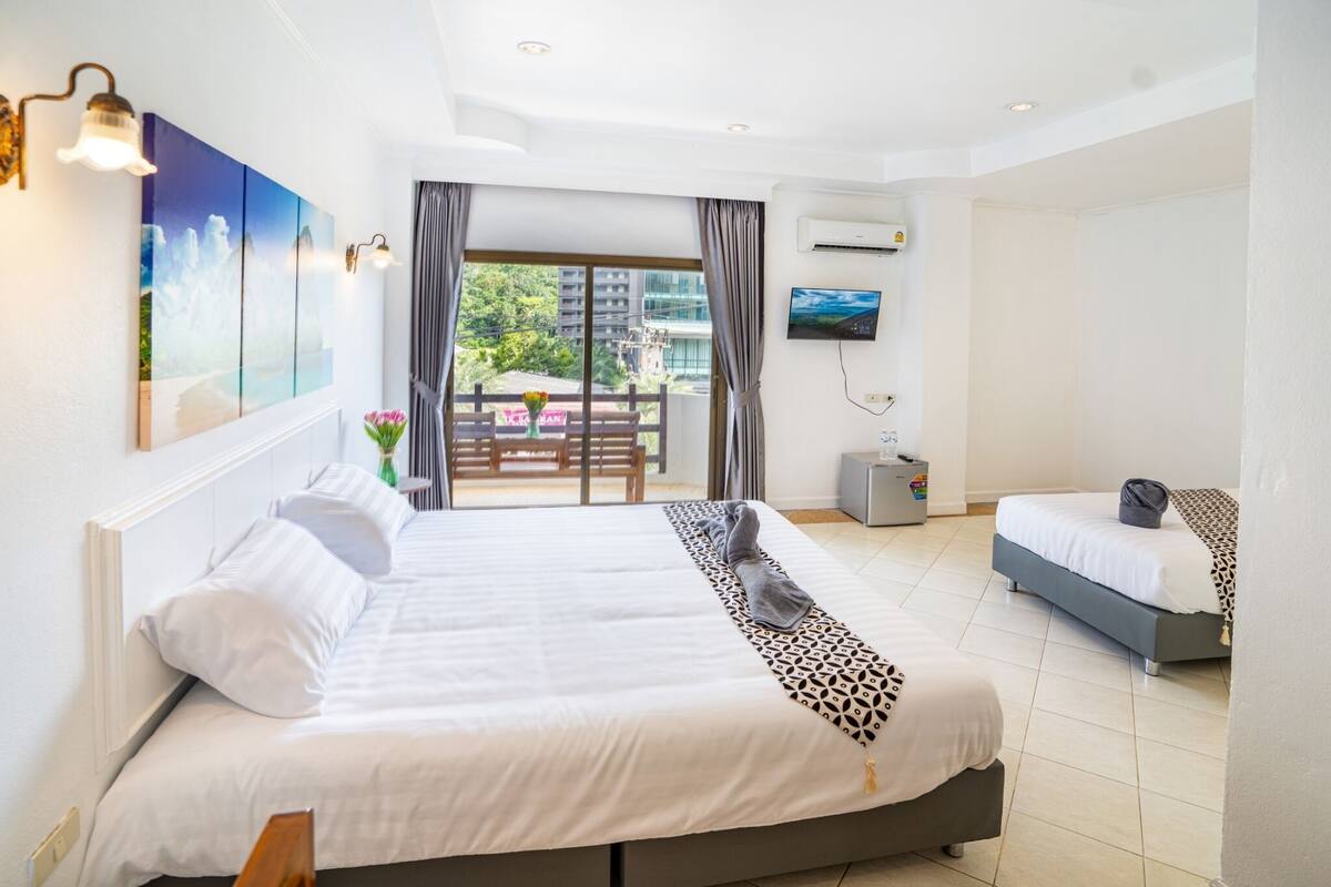 Ein Hotelzimmer mit einem großen Bett, einem kleineren Bett, einem Fernseher und einem Balkon mit Blick auf Gebäude.