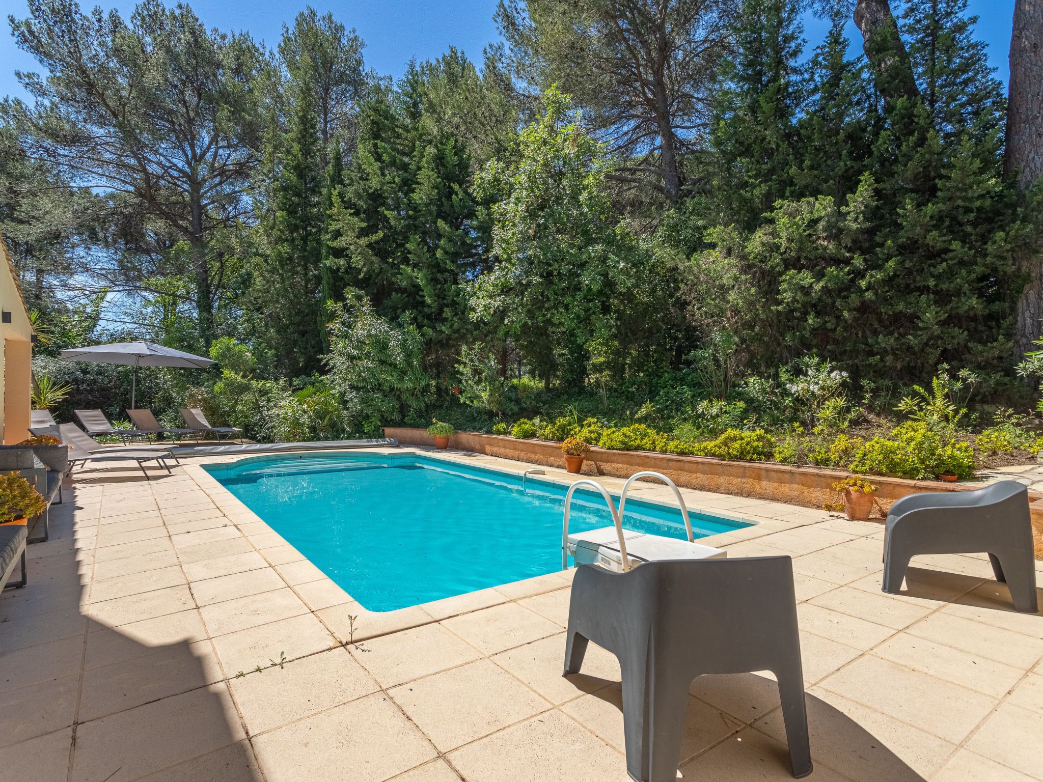 Une piscine rectangulaire avec un escalier, entourée d’une terrasse carrelée. On y trouve des chaises longues et une petite table avec un parasol.