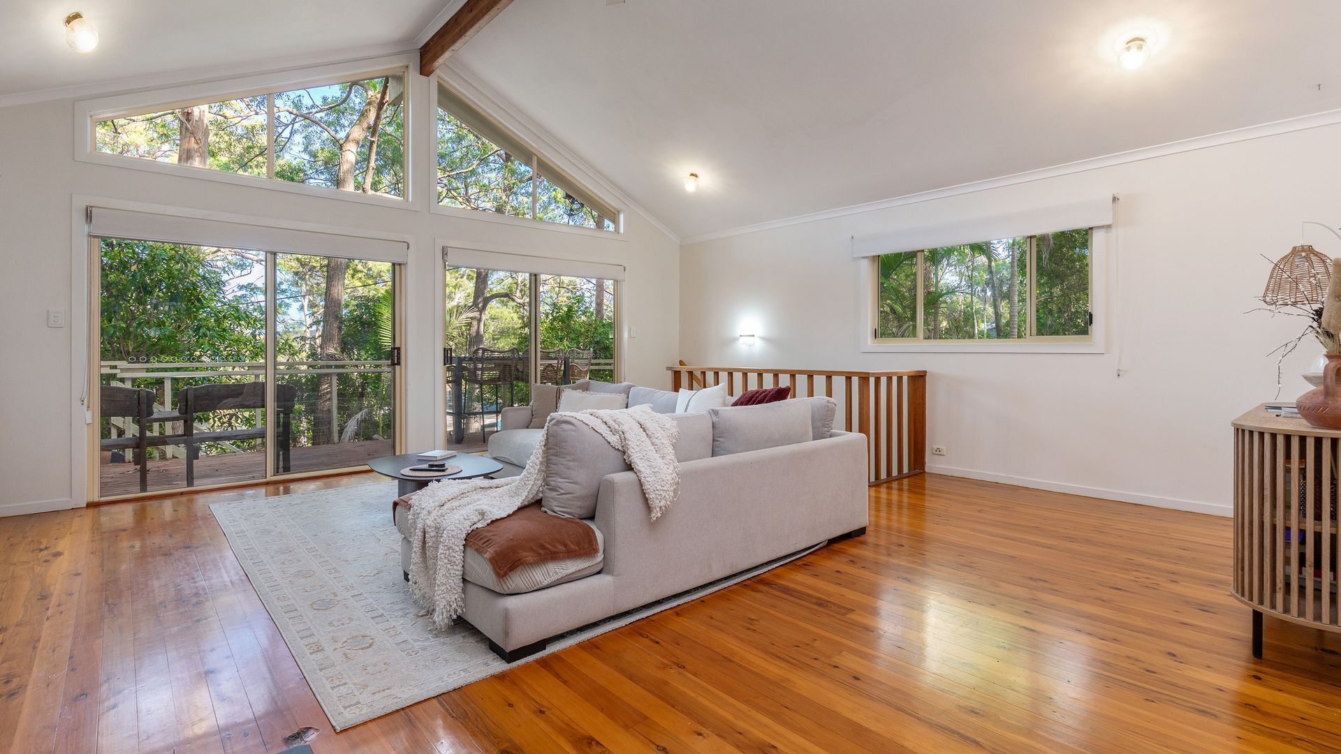 A spacious living room with a large sofa, wooden flooring, and large windows offering views of trees.
