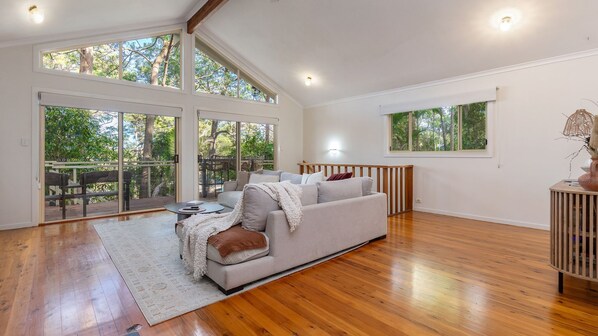 A spacious living room with a large sofa, wooden flooring, and large windows offering views of trees.