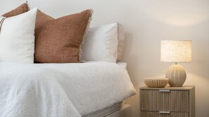 A neatly made bed with white bedding and two pillows, one brown and one white. Beside the bed is a small wooden nightstand with a lamp and two decorative bowls.