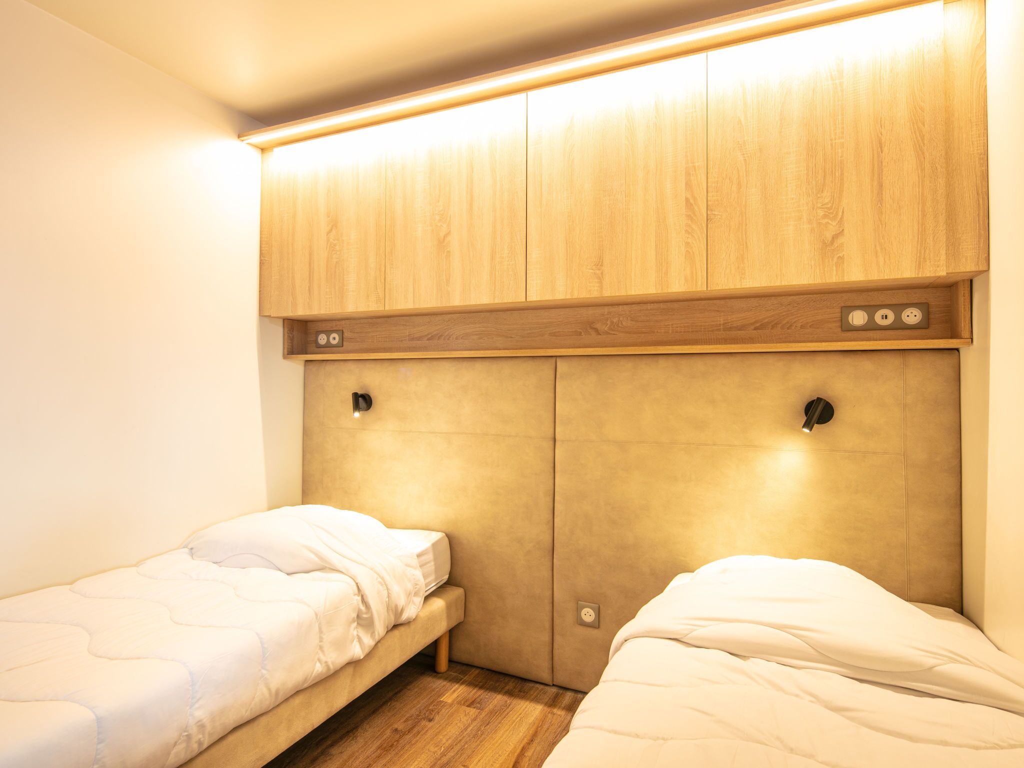 A bedroom with two single beds, wooden headboard, built-in lighting, and a small shelf with electrical outlets.