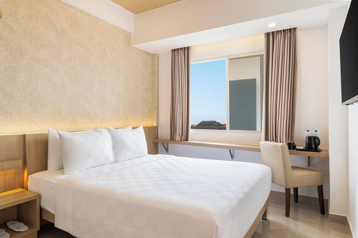 Ein Hotelzimmer mit einem großen Bett, einem Schreibtisch mit Stuhl, einem Fenster mit Vorhängen und einem an der Wand befestigten Fernseher.