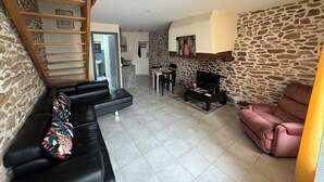 Un salon avec un mur en pierre, une cheminée, un canapé noir, un fauteuil rose et un escalier en bois.
