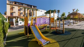 Un parque infantil con un tobogán y estructuras para escalar, rodeado por un edificio con balcones y una palmera.