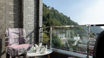 A balcony with a stone wall, a cushioned chair, a small table with cups and a coffee pot, and a view of a hillside with buildings.