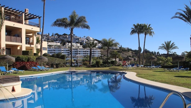 A swimming pool with lounge chairs, palm trees, and a building with balconies in the background. - Garden Apt In Los Pinos, Los Arqueros Golf (Benahavis)