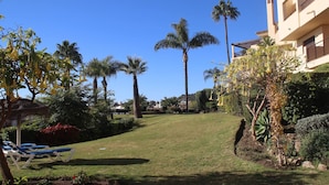 A well-maintained garden with palm trees, a grassy slope, and lounge chairs. - Garden Apt In Los Pinos, Los Arqueros Golf (Benahavis)