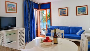 A living room with a TV, a blue sofa, a dining table with a bowl of fruit and a bottle of wine, and a view of palm trees through a window. - Playa D Las Américas-casa Rucaba (Adeje)