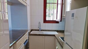 A compact kitchen with white cabinets, a stainless steel refrigerator, a built-in oven, and a window with a red frame. - Playa D Las Américas-casa Rucaba (Adeje)