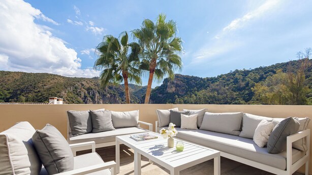 A terrace with a white sofa set, a table, and a view of a mountainous landscape with palm trees. - El Casar Townhouse Benahavis (Benahavis)