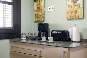 En køkkenbordplade med kaffemaskine, brødrister og papirhåndklædeholder. To krukker med låg og et vindue med skodder.