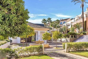 A courtyard with a fountain, palm trees, and a building with a tiled roof. - El Casar Townhouse Benahavis (Benahavis)