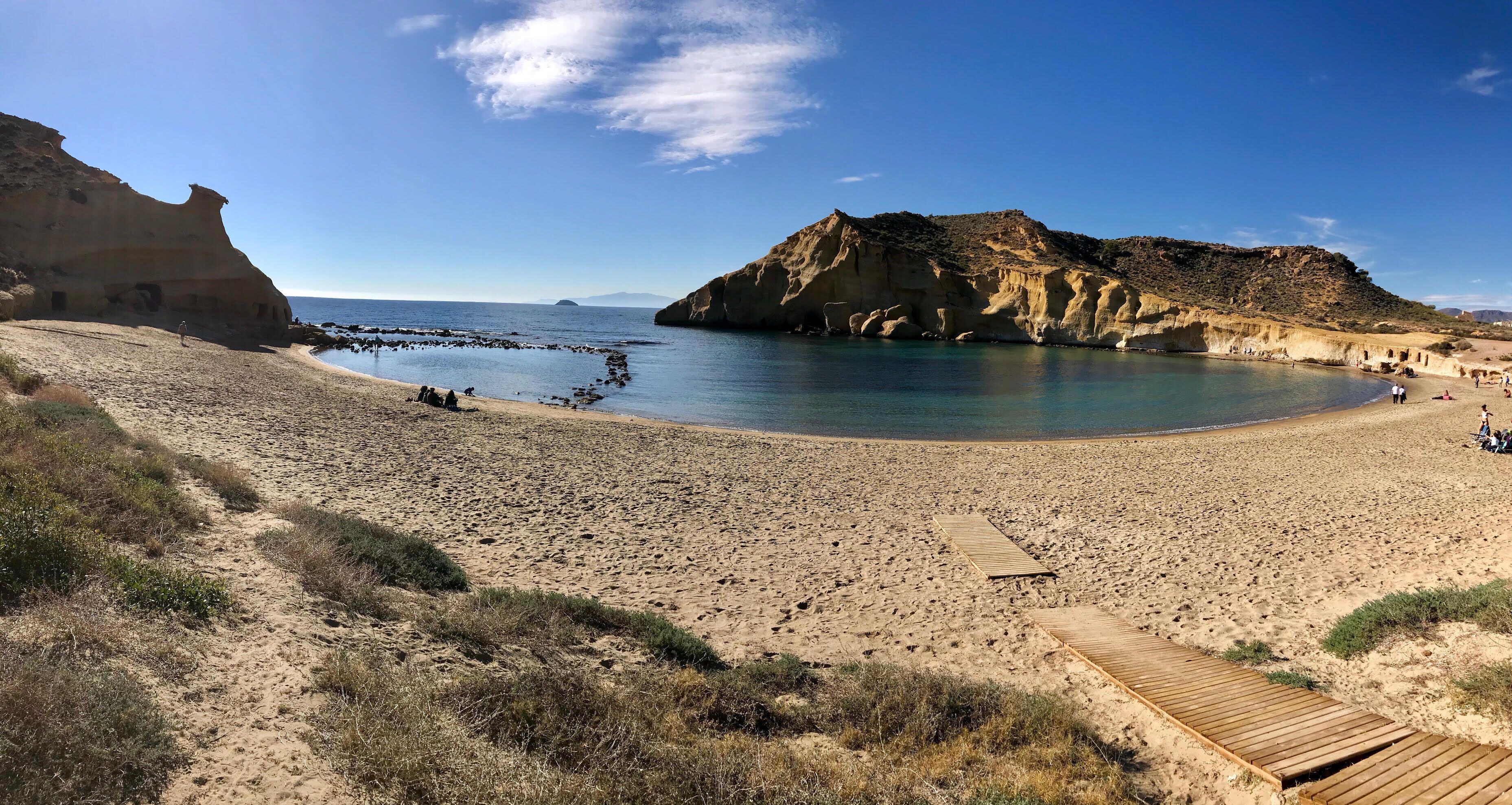 Una platja amb una passera de fusta que condueix a l'aigua, envoltada de dunes sorrenques i penya-segats rocosos.