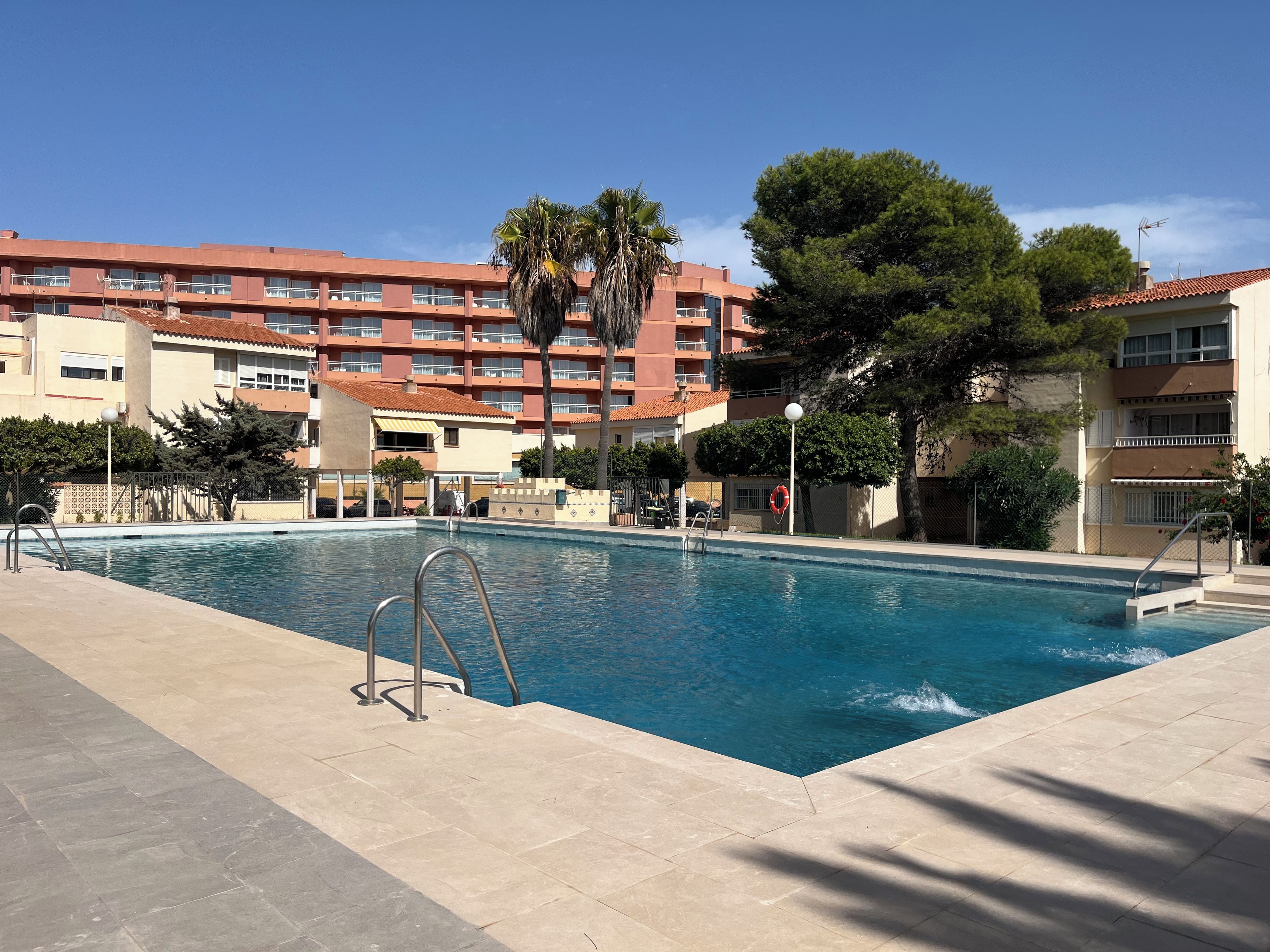 A swimming pool with a metal ladder, surrounded by a tiled deck, with buildings in the background.